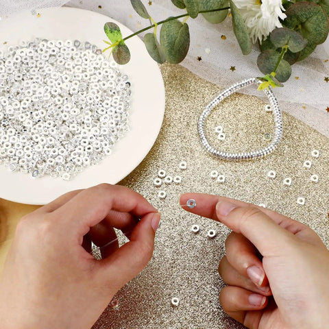 Hands holding six millimeter flat round spacer beads pack of 1500 silver rondelle beads for jewelry making on gold surface with greenery.