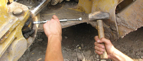 Grease joint rejuvenator tool in use on yellow heavy equipment; hands hold a metal rod and wooden hammer.
