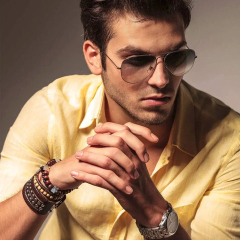 Man wearing sunglasses and wrist bracelets with a thoughtful expression and a yellow shirt in a moody lighting setup.