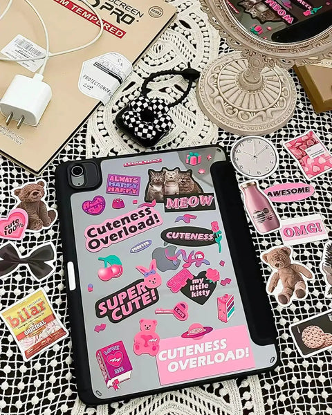 Tablet cover decorated with cute pink and black stickers on lace table, surrounded by items like charger and small mirror.