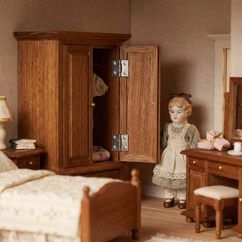 Miniature bedroom scene with dollhouse furniture, showcasing wooden wardrobe with metal hinges and a doll standing nearby.