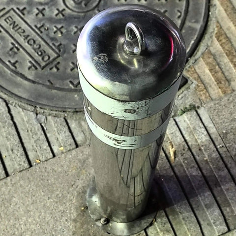 Stainless steel street bollard with lifting ring cap next to manhole cover in urban pavement setting.