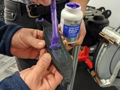 Permatex Ceramic Extreme Brake Parts Lubricant bottle (8 oz) with purple lubricant on brake pad