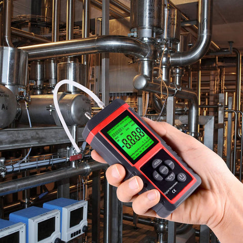 Handheld digital manometer with red and black case and green LCD display measuring HVAC air pressure via a white hose.