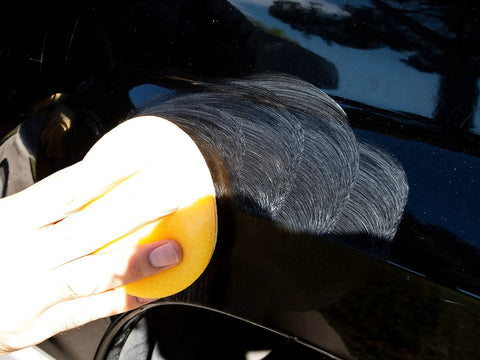Person using Meguiars ScratchX 7 Oz Liquid on a black car panel to remove scratches with an applicator pad.
