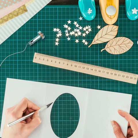 Crafting table with cutting tools, cutting mat, ruler, and hands holding a craft knife with paper, scissors, and embellishments visible.