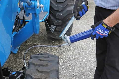 Lever-action grease gun with 18-inch whip hose attached to a blue tractor, gloved hand guiding.