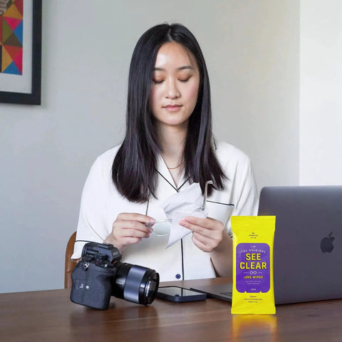 Woman using Lens Cleaning Wipes For Glasses And Screens Streak Free Scratch Free 192 Wipes on glasses near laptop and camera.