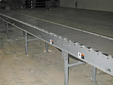 Long industrial conveyor belt in a warehouse setting with metal support legs and bolts.
