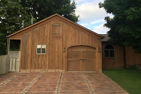 Wooden barn with a brick driveway, featuring an arched wooden gate door, surrounded by trees on a sunny day.