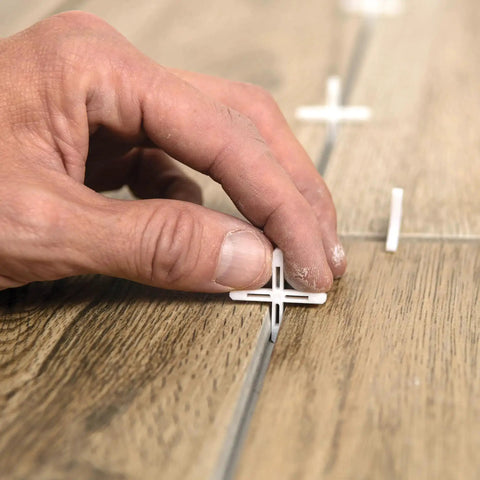 Hand placing 1/8 Inch Hard Tile Spacers for Traditional or Leave-In Installations, 300 Pack, ensuring consistent grout joints on wood-look tiles.