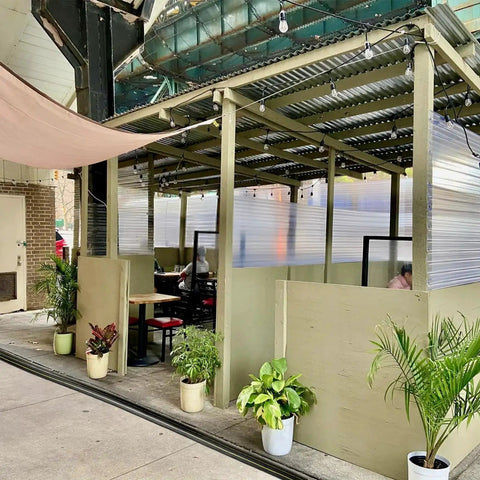Outdoor seating area with corrugated roof, potted plants, and semi-private dining booths on a sidewalk.