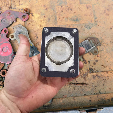 Rectangular black automotive gasket with circular metal insert and four corner bolts, held in hand
