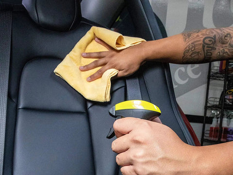 Person using a yellow spray bottle on a car seat, wiping with a microfiber cloth for cleaning and detailing.