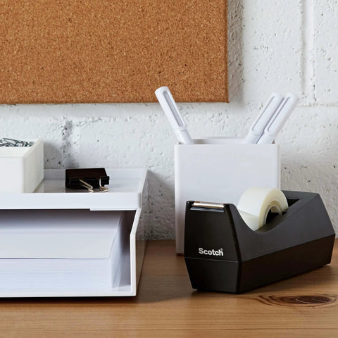 Black desktop tape dispenser, weighted base, next to desk organizer and pens, single unit, for one-hand dispensing, refillable 1-in core.
