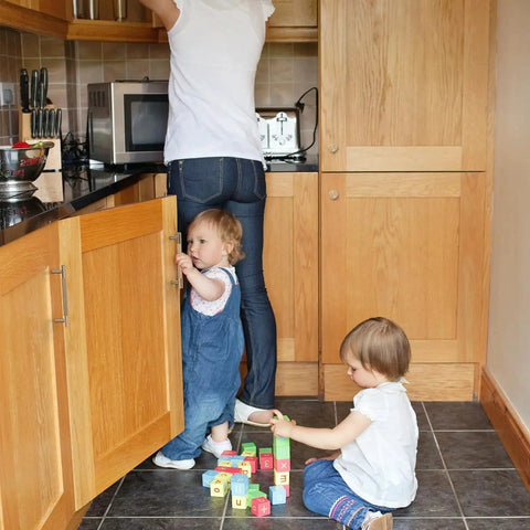 Two toddlers playing with blocks in a kitchen, woman reaching into a cupboard above, wooden cabinets with silver handles visible.