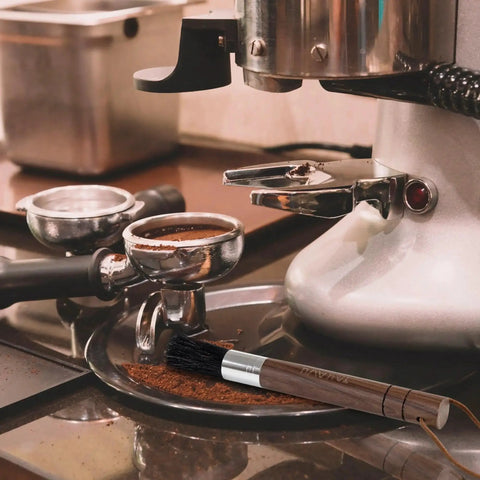 Takavu coffee grinder cleaning brush with wooden handle and black boar bristles beside a stainless espresso machine.
