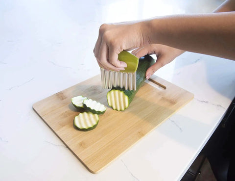 Person using Crinkle Cutter Knife With Wavy Edge For Fries And Veggies Kids Friendly Kitchen Tool Stainless Steel BPA Free Colors May Vary Great For Fun Cooking And Holiday Stocking Stuffers Joie to slice cucumber on cutting board.