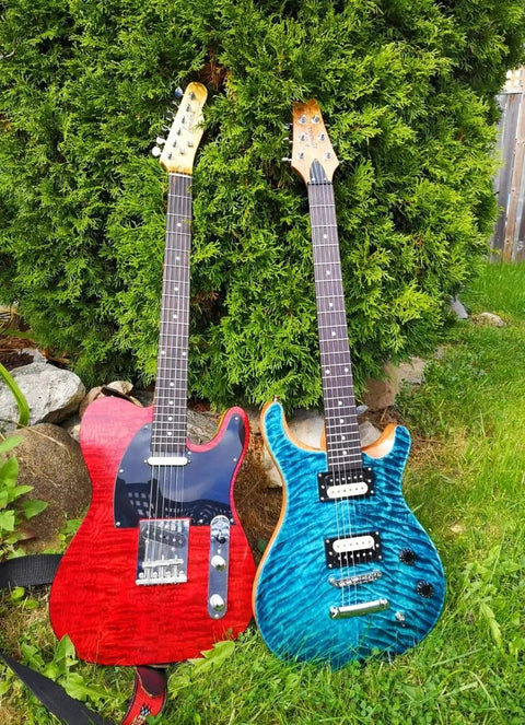 Two vibrant electric guitars, one red and one blue, leaning against a green bush outdoors, showcasing wood grain and body design.