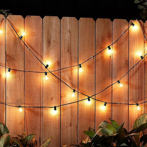 Outdoor string lights hanging on a wooden fence, illuminated at night with warm bulbs, surrounded by plants below.
