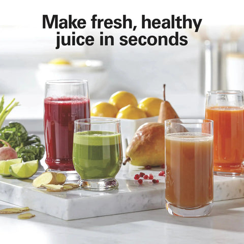 Juicer machine on kitchen counter with four glasses of colorful juice (red, green, orange) and fruit in background.