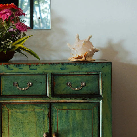 Green cabinet with Six Pack Vintage Drop Bail Cabinet Handles In Antique Brass Finish, displayed with flowers and conch shell decor.