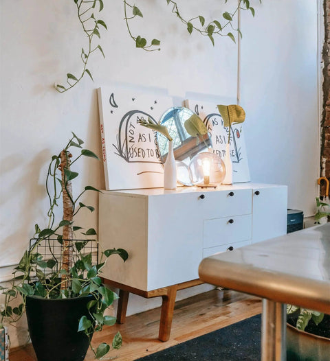Modern white cabinet surrounded by green plants, featuring decor and lighting on top, minimal home interior design.