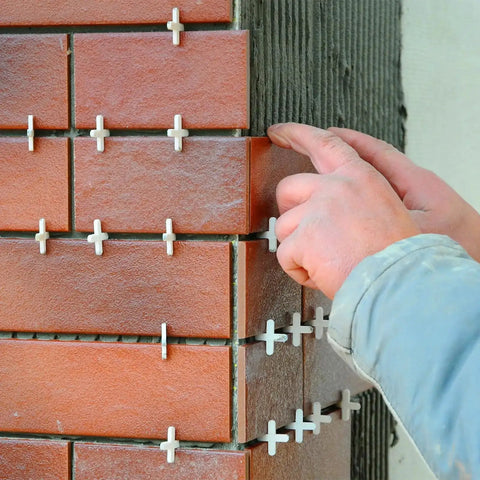 Hands placing 1200 Pcs Cross Shape Tile Spacers 1mm Gap For Ceramic Tile Installation, ensuring precise grout lines between red tiles on a wall.