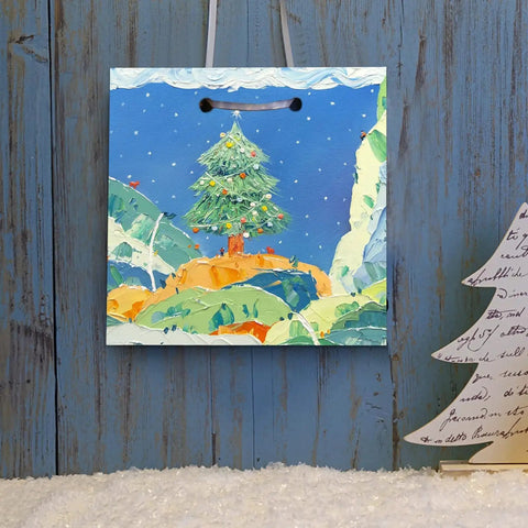 Gift bag with painted Christmas tree, mountains, and snow; wooden background and paper-craft tree visible.