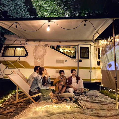 People sitting outside an RV watching a movie projected on a screen at night, surrounded by string lights and blankets.