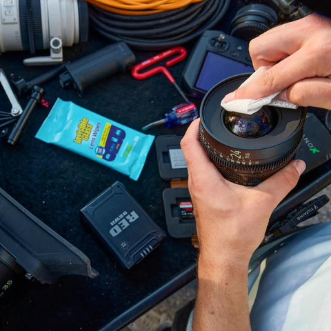 Person using Streak Free Lens Cleaning Wipes For Glasses Cameras Binoculars 16 Count on a camera lens with electronics on a table