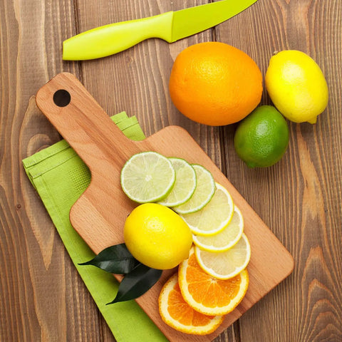 Wooden cutting board with sliced lemons, limes, and oranges next to whole citrus fruits and a green knife on a wooden surface.