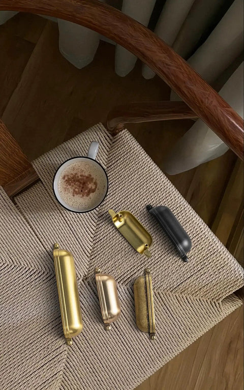 Assorted metallic drawer pulls on woven chair with coffee mug, showing different finishes and styles.