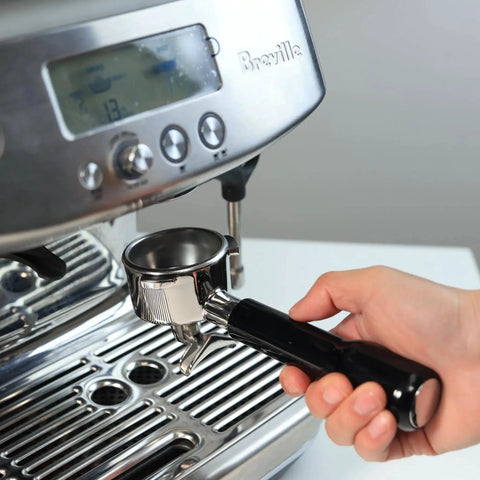 Close-up of chrome 54mm double-spout portafilter with black ABS handle held next to a Breville espresso machine.