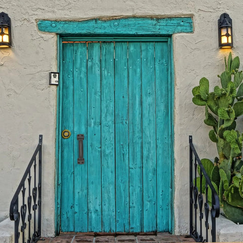 Turquoise wooden door with iron barn door pull handle, surrounded by cacti and lanterns.