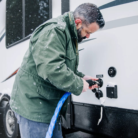 Camco 25-ft heated RV water hose connected to a trailer water inlet in snow; blue hose and user in green jacket.
