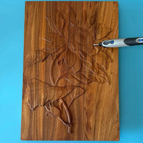 Wooden board with a carved flower design, being detailed with a rotary tool on a blue background.