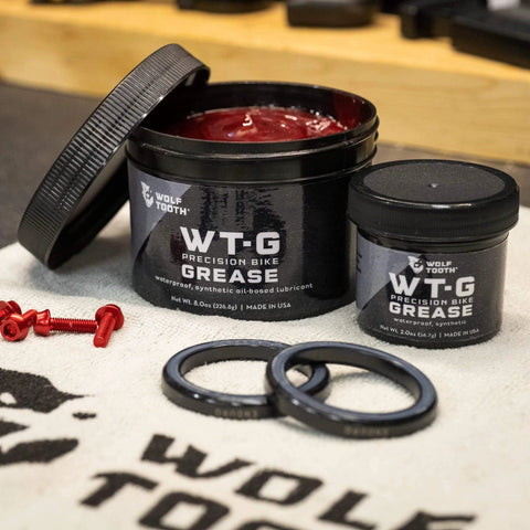 Wolf Tooth WT-G Precision Bike Grease containers, 2 oz and 8 oz, on a workshop table with components visible.