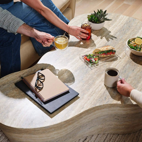 Marble coffee table with notebooks, glasses, drinks, salad, and sandwich with people reaching for beverages.