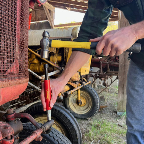 Grease Buster hand tool with red handle used on a tractor joint; yellow hammer nearby