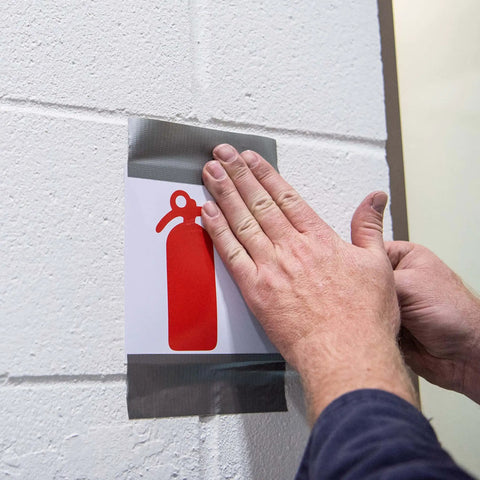 Person applying Value Thin Silver Duct Tape Single Roll to secure a fire extinguisher sign on a wall.
