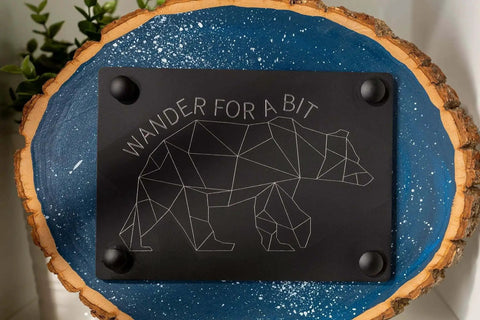 Wall art with geometric bear design and 'Wander For A Bit' text on a black plaque mounted on blue and wood background.