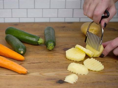 Crinkle Cutter For Fruits And Veggies With Non Slip Comfort Rubber Handle And Zigzag Stainless Steel Blade, Endurance Collection By RSVP International, 18 8 Stainless Steel, Dishwasher Safe, 4.5 By 3 Inches cutting potatoes on a wooden surface with carrots and zucchini nearby.