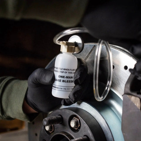White ABN One-Man brake bleeder bottle with magnet, held by gloved hand beside a brake rotor and caliper.