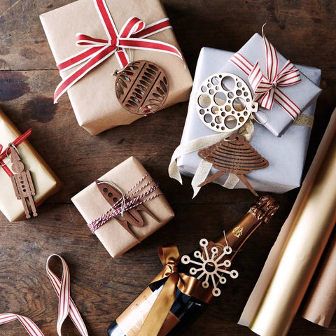 Assorted gift boxes with ribbons and wooden ornaments, a bottle of champagne, and wrapping paper on a wooden surface.