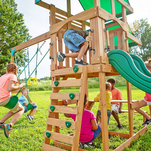 Children playing on a wooden playset with swings and slide in a grassy yard