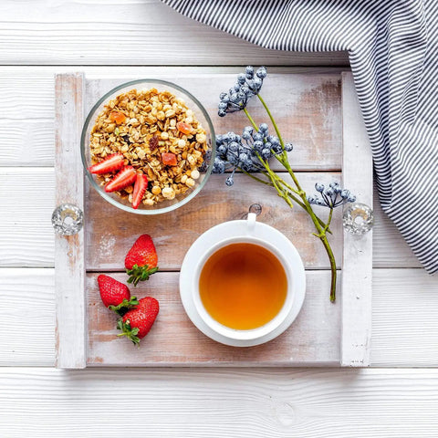 Breakfast tray with granola and strawberries, tea cup, and purple flowers on a wooden tray with diamond knobs on a striped cloth.