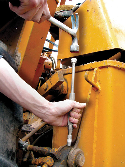 Grease joint rejuvenator kit tool; silver metal rod held by hand against yellow industrial machinery.