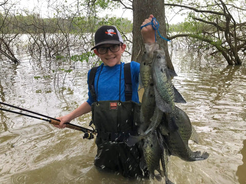 Person holding a catch of fish in a flooded forest, wearing a cap and waders, showcasing successful use of fishing tackle kit.