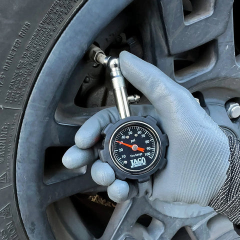 JACO Deluxe Tire Pressure Gauge, 100 PSI, silver gauge with black dial, measuring car tire valve with gloved hand